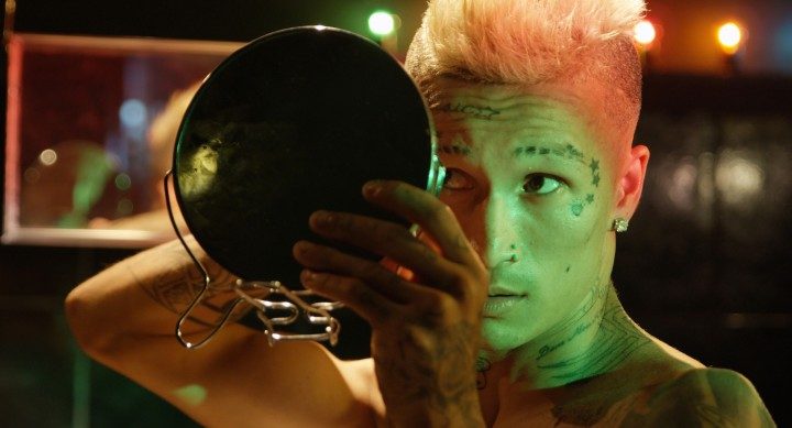 Junge Person mit tätowierter Haut, die sich in einem Spiegel betrachtet, umgeben von buntem Licht.